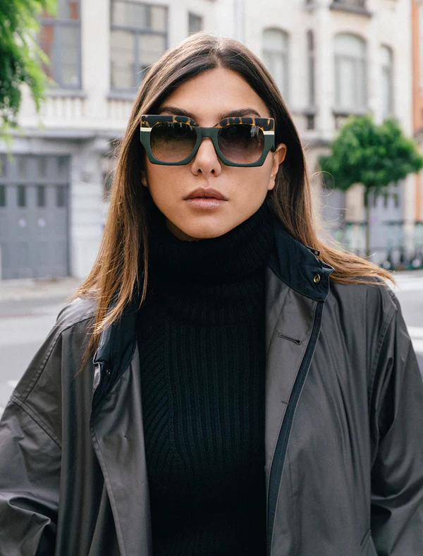 Mulher com óculos de sol quadrados verde escuro com padrão animal print nas hastes, casaco cinza e camisola preta em rua urbana