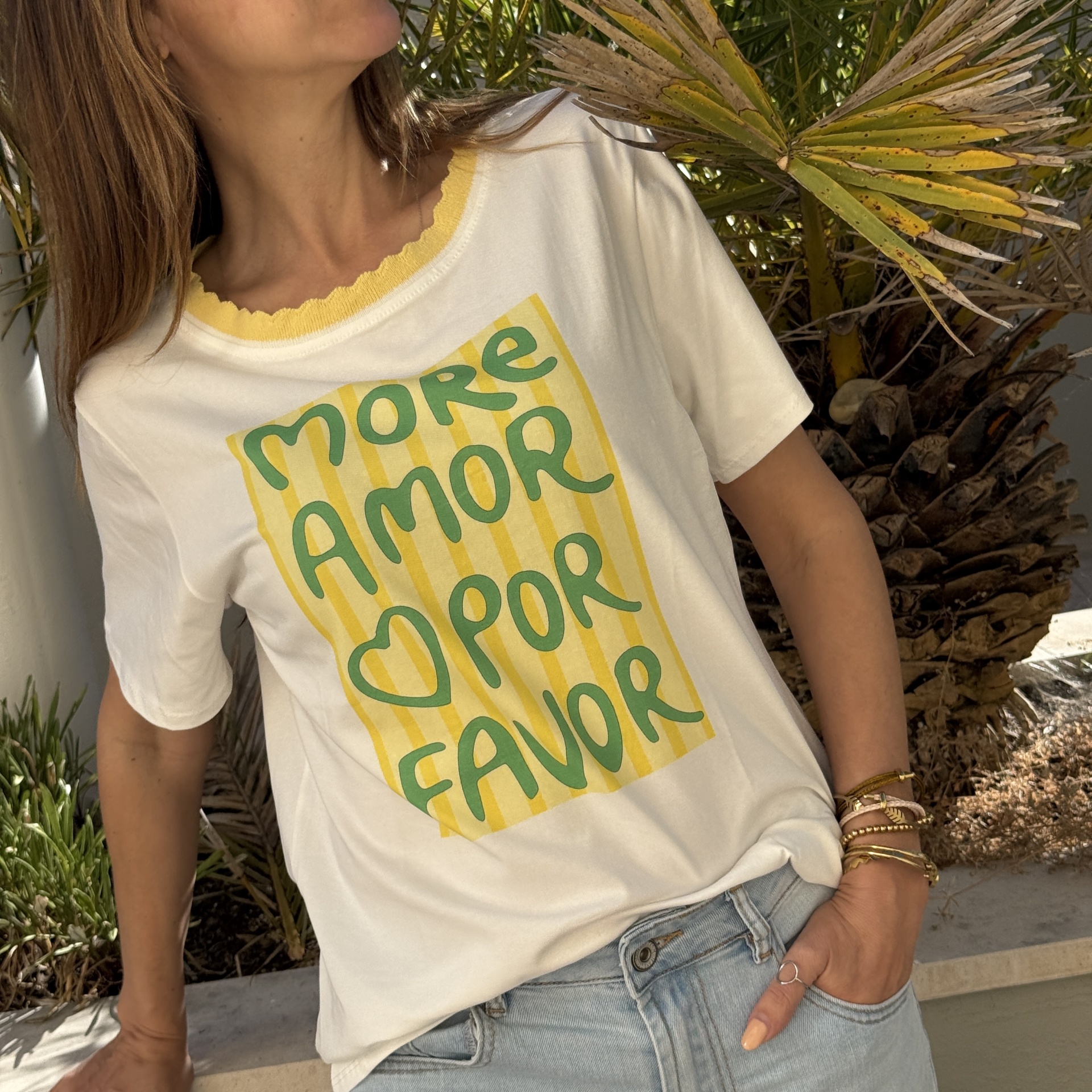 T-shirt branca com gola amarela canelada e texto verde em riscas amarelas, usada por mulher junto a plantas.