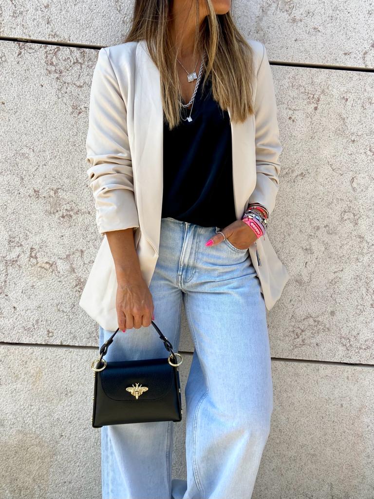 Mulher com blazer creme, t-shirt preta e jeans claros segurando bolsa preta pequena com detalhe dourado.