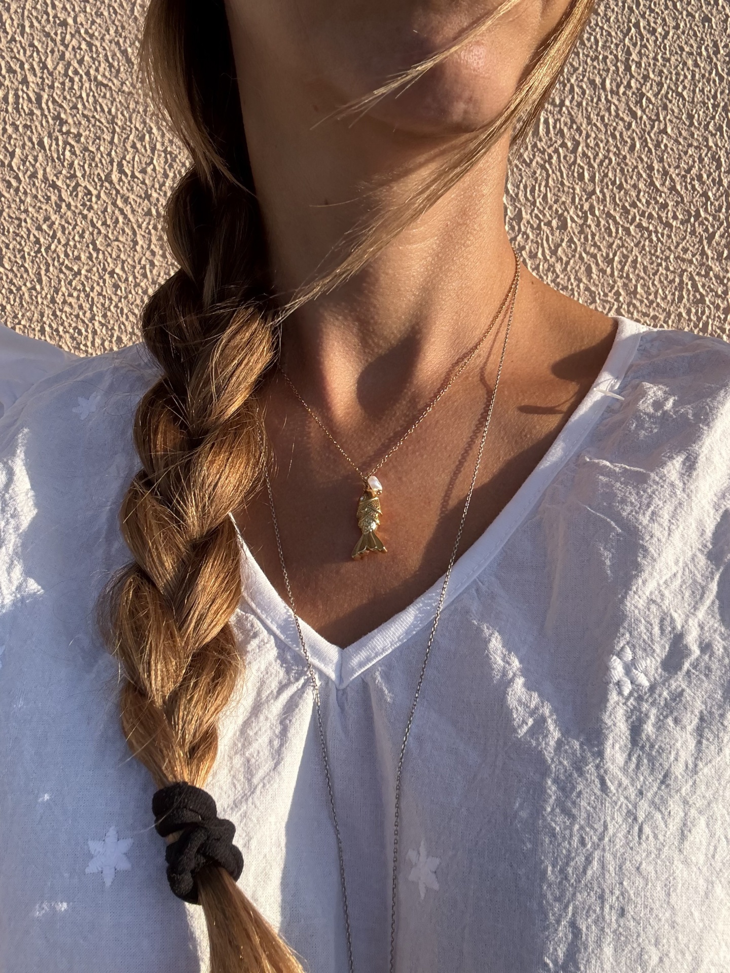 Colar dourado com pendente de figura humana e pérolas usado com camiseta branca de tecido leve e cabelo loiro em trança.