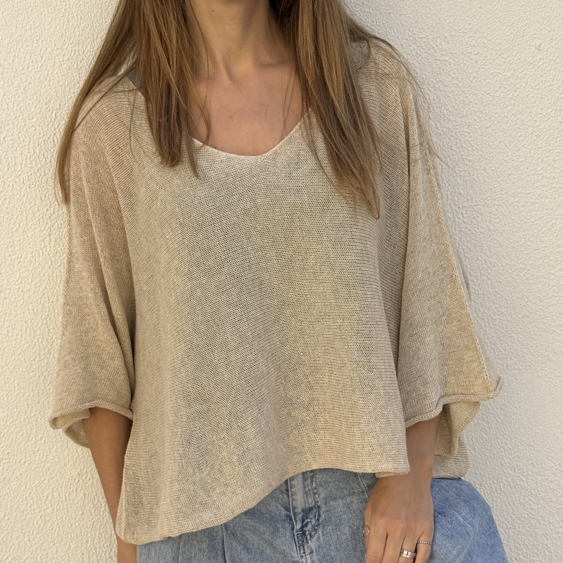 Blusa bege de malha com decote redondo e mangas largas cortadas junto a parede branca