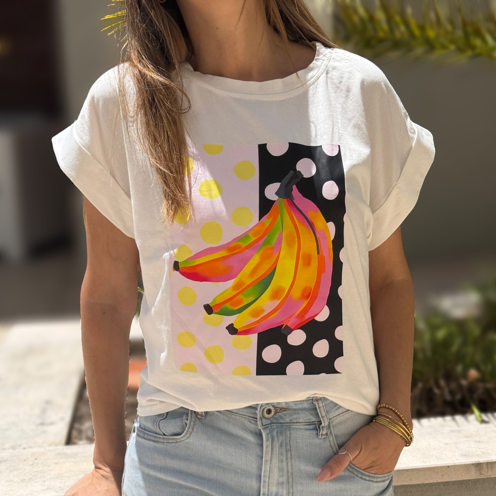 Homem a usar t-shirt branca com estampa de bananas coloridas e bolinhas, combinada com jeans claros e pulseiras douradas