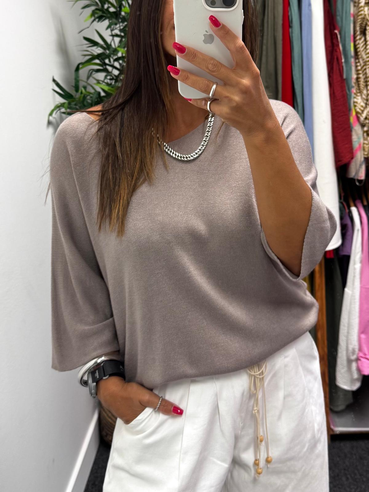 Pessoa em frente ao espelho com camisola bege e calças brancas num cenário de loja com roupa pendurada.