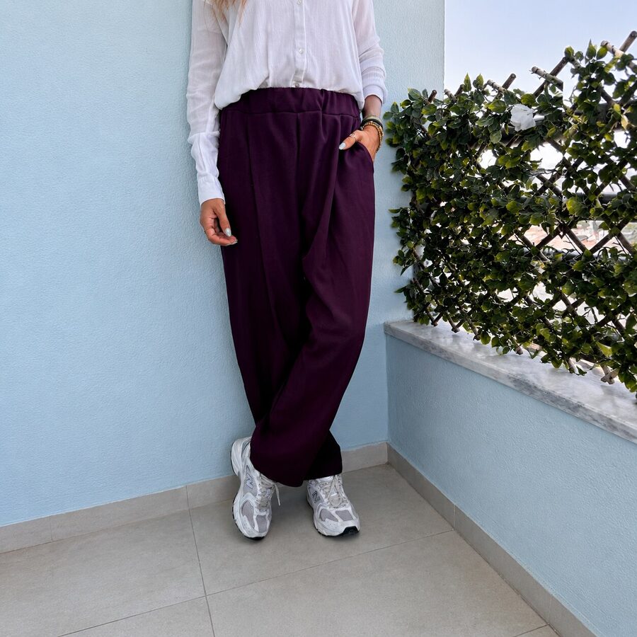 Pessoa com calças roxas largas, camisa branca e ténis brancos junto a parede azul clara e grade com plantas