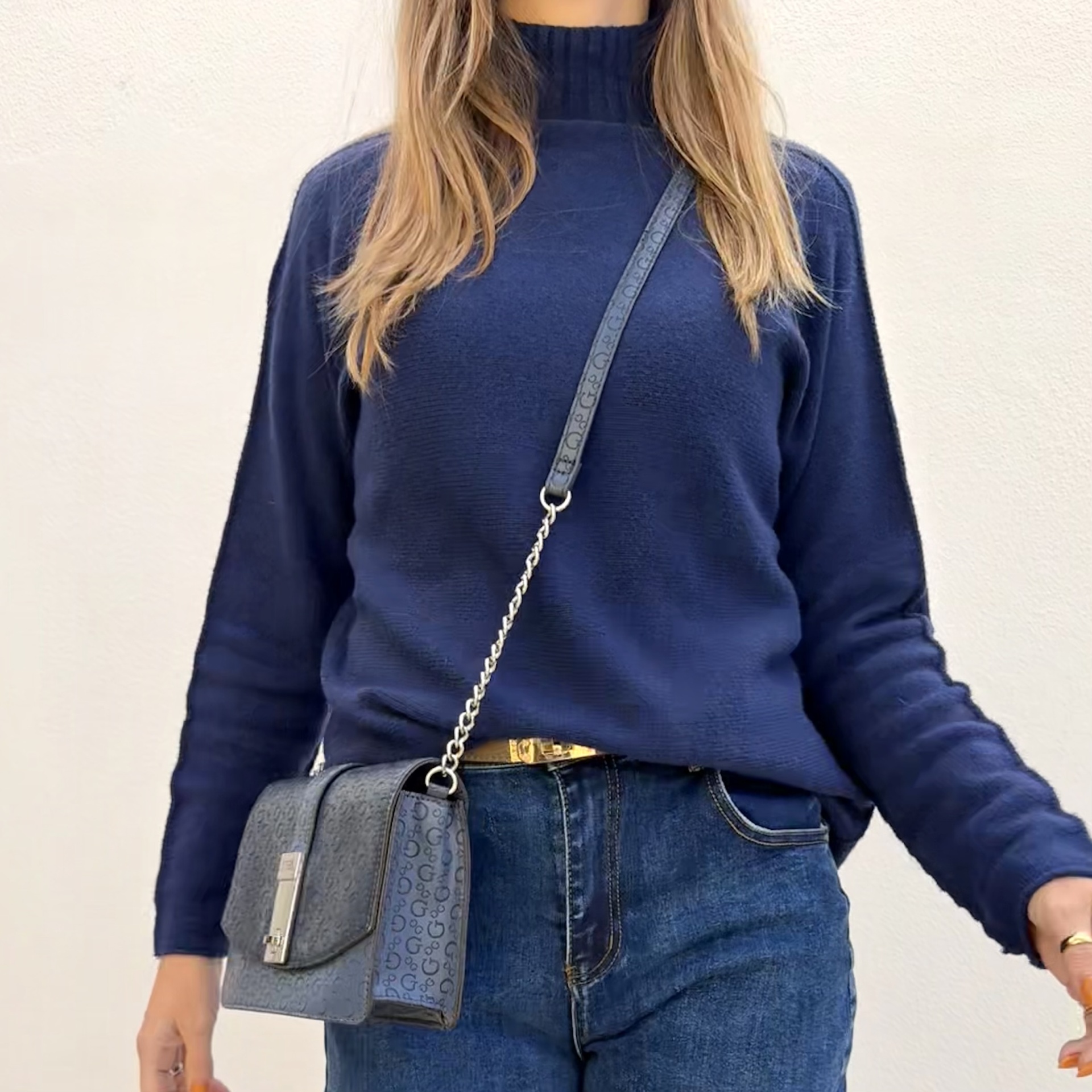 Mulher com camisola azul escura, calças de ganga azuis e bolsa tiracolo cinzenta com padrão de letras.