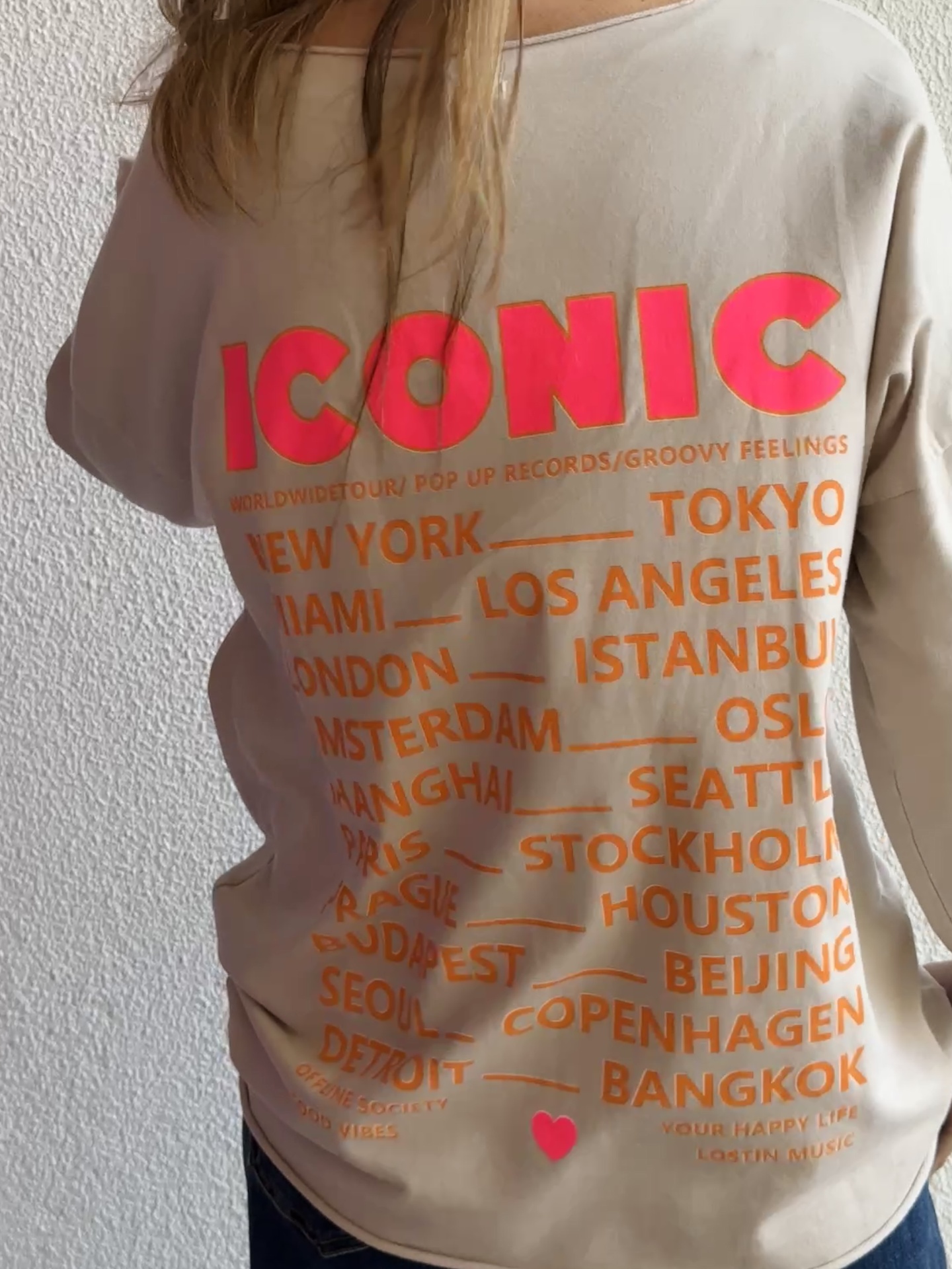 T-shirt bege com texto em vermelho e laranja nas costas, incluindo ICONIC e nomes de cidades.