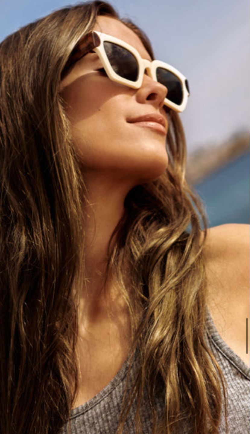 Mulher a usar óculos de sol brancos e castanhos com camisola cinza ao ar livre junto ao mar