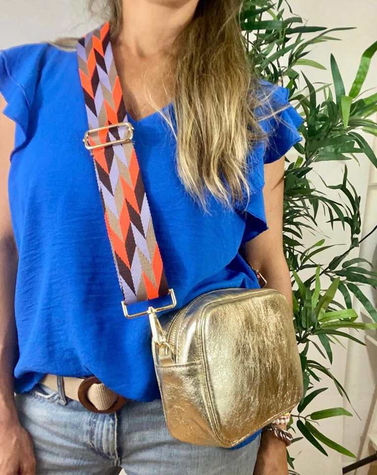 Bolsa quadrada metálica dourada com correia estampada colorida, roupa azul e jeans, planta verde ao fundo