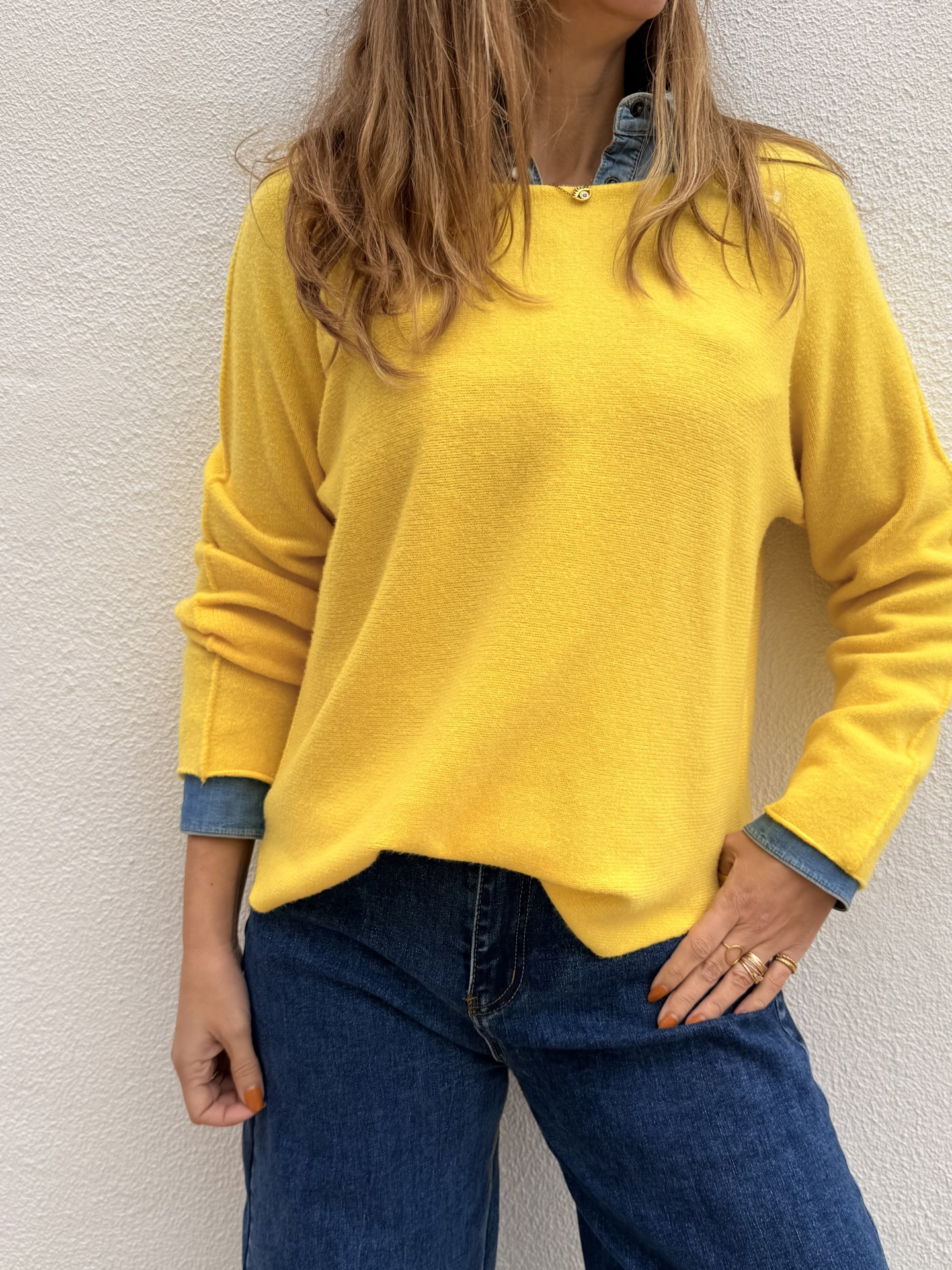 Pessoa com pullover amarelo e camisa de ganga azul, calças de ganga azul escuro, contra parede branca.