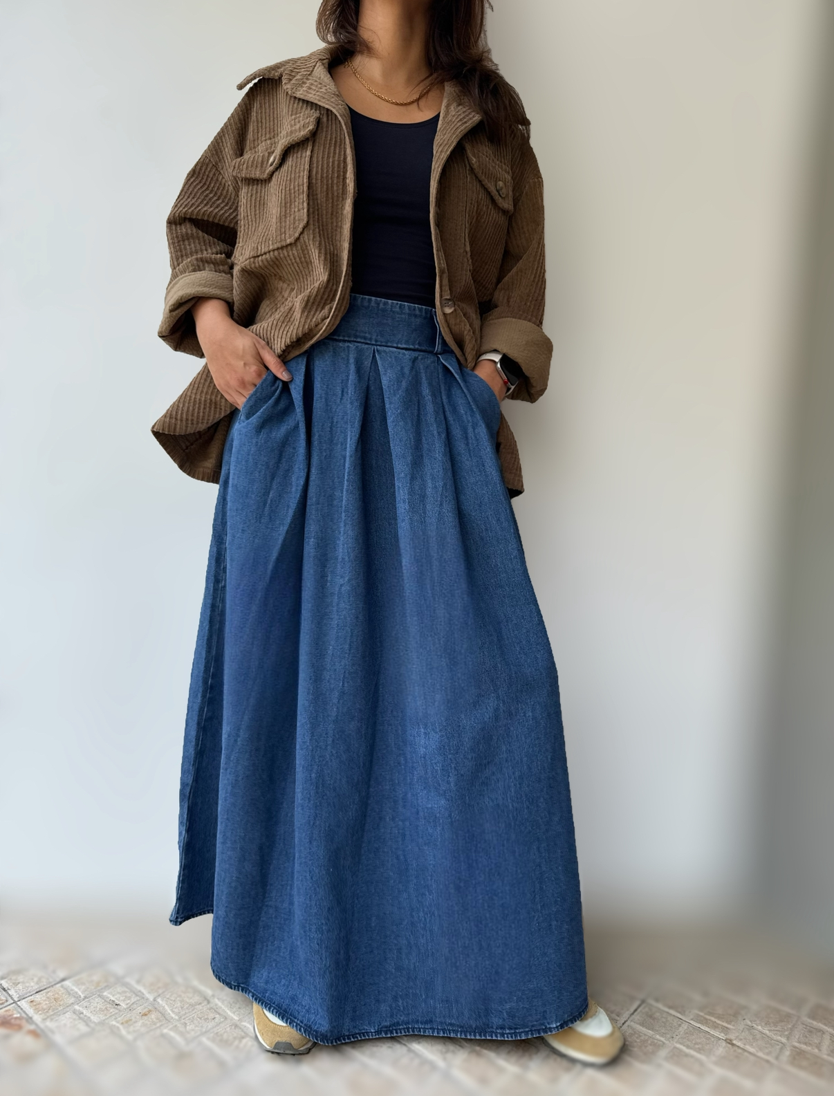 mulher a usar saia de ganga azul comprida, top preto e camisa castanha de veludo cotelê