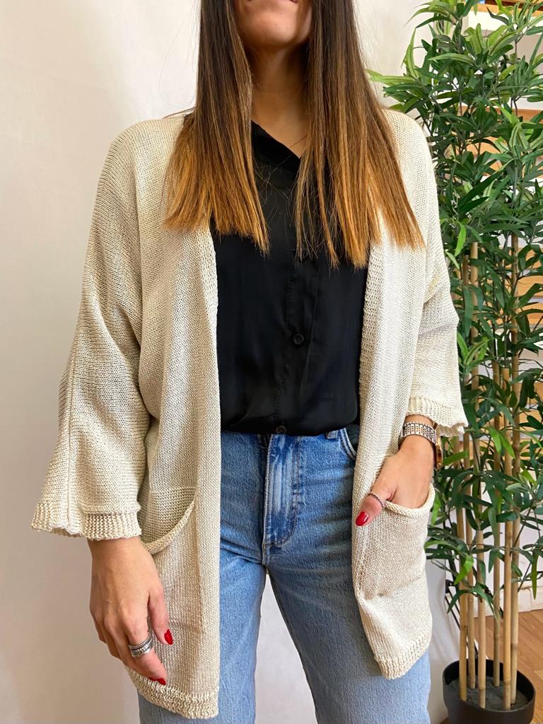 Mulher veste casaco aberto bege de malha com bolsos, camisa preta e jeans azul claro junto a planta em vaso