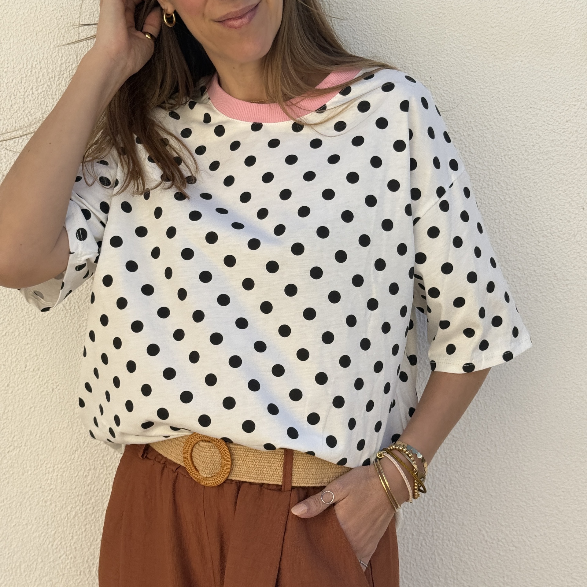 Mulher com t-shirt branca de bolinhas pretas e gola rosa, calças castanhas e cinto de tecido natural