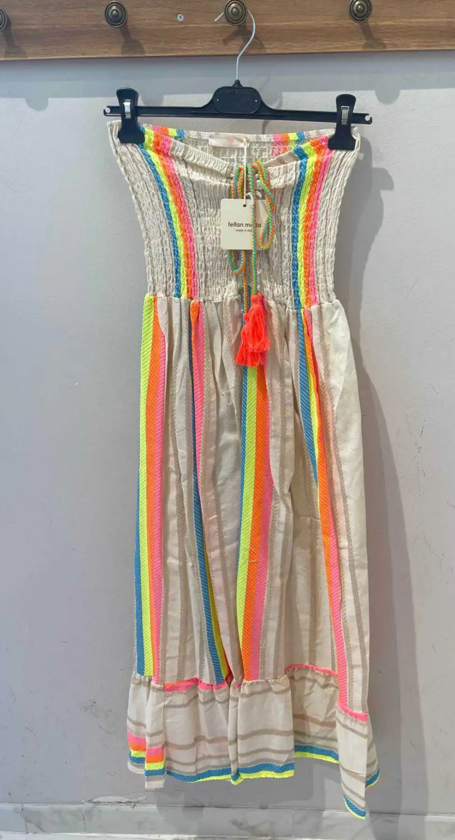 Vestido longo bege com riscas coloridas e cinto com pompons cor-de-rosa num cabide preto