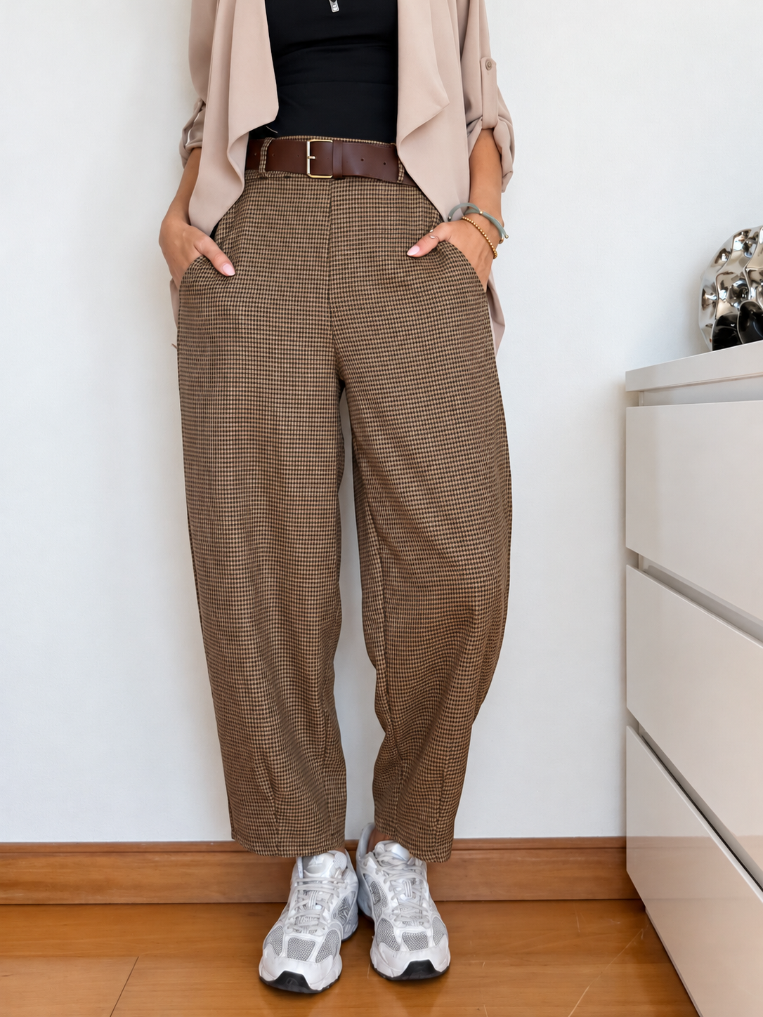 Calças xadrez castanhas com cinto castanho, t-shirt preta, blazer bege e sapatilhas brancas num ambiente interior
