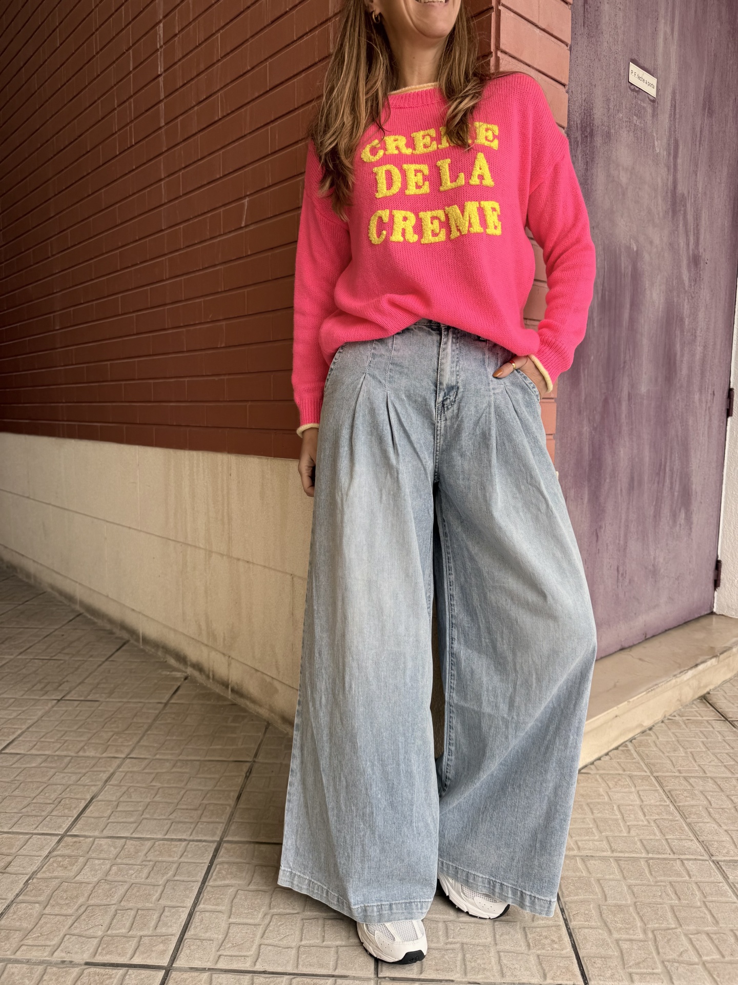 Pessoa com sweatshirt rosa e calças largas de ganga azul clara, com ténis brancos, numa rua de chão de ladrilhos e paredes vermelha e roxa.