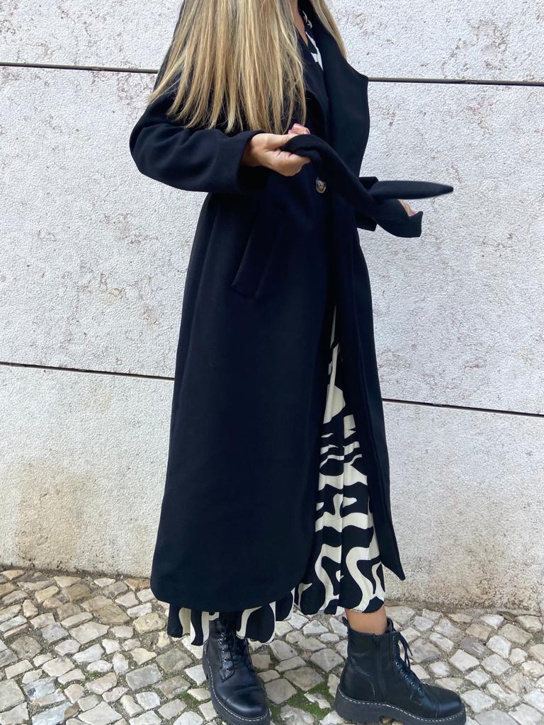 Casaco comprido preto com vestido padrão abstrato preto e branco e botas pretas no chão de calçada portuguesa