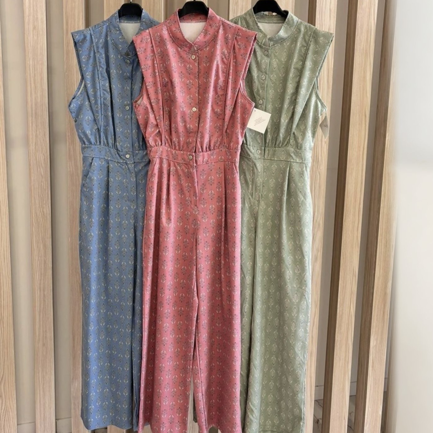 Três macacões estampados em azul, rosa e verde claro pendurados em cabides numa estrutura de madeira.