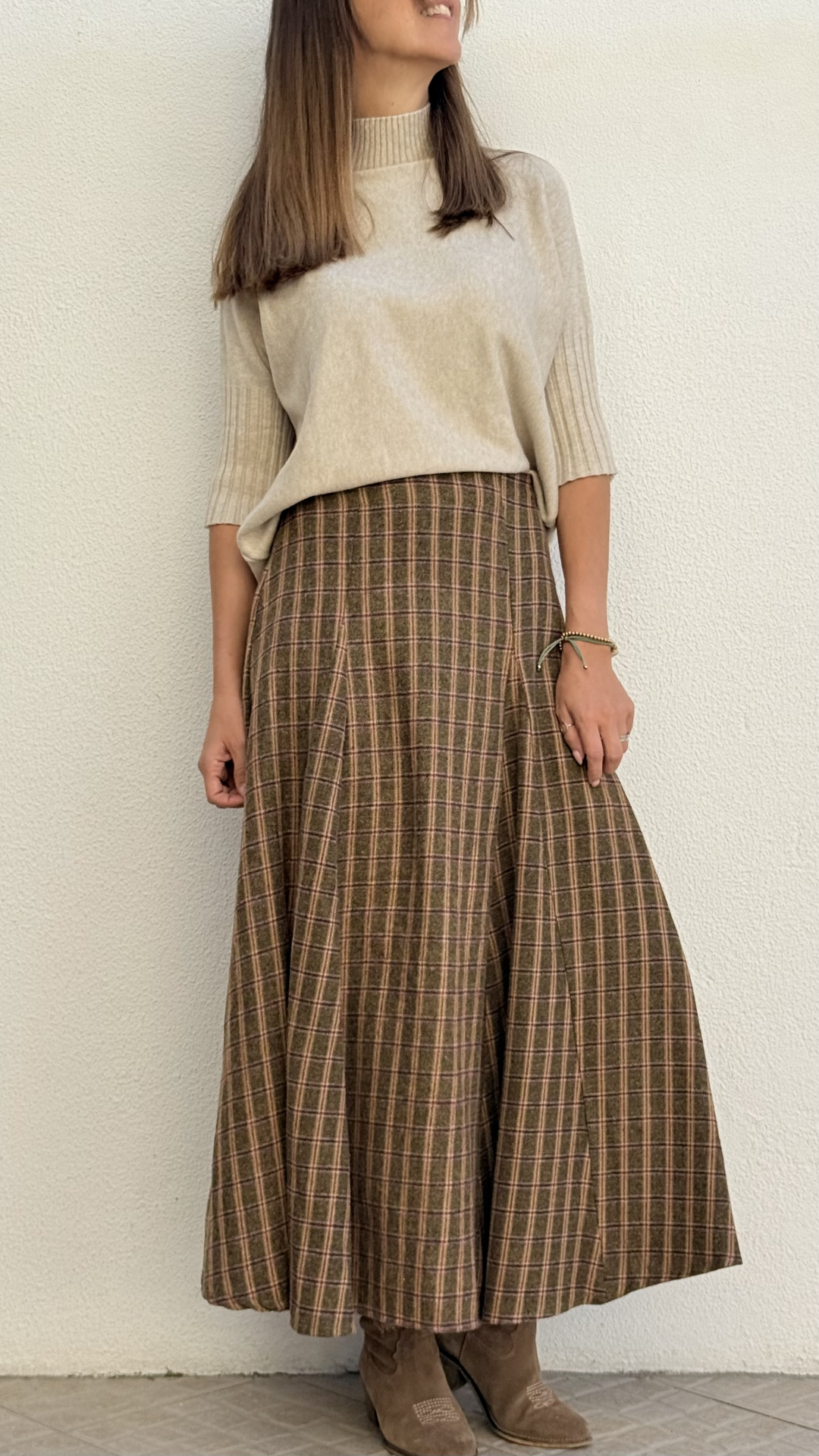 Mulher com camisola bege, saia xadrez castanha e botas castanhas em fundo branco.