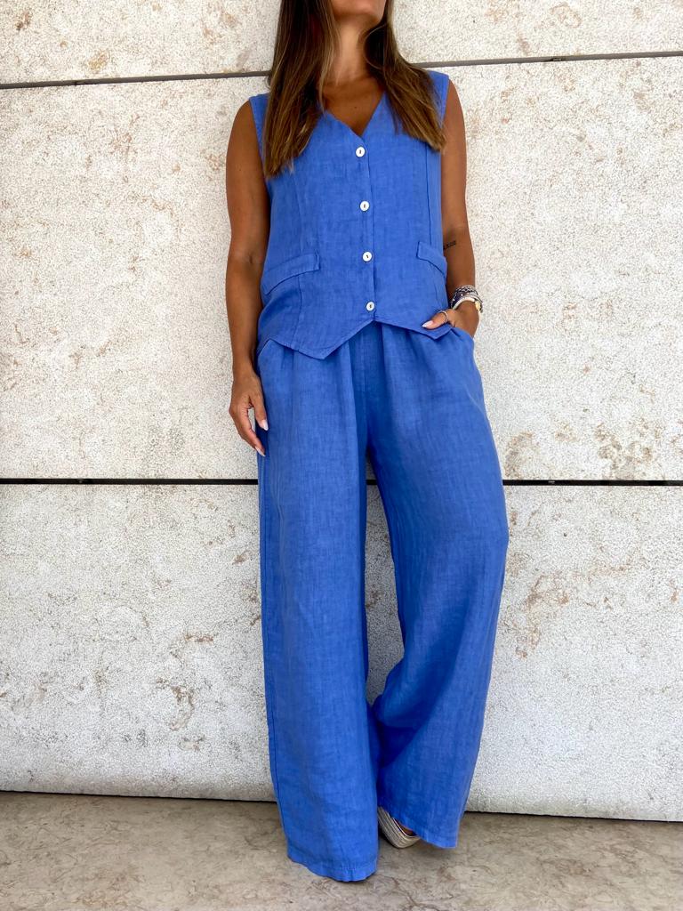 Conjunto feminino azul de colete e calças largas junto a parede de pedra clara