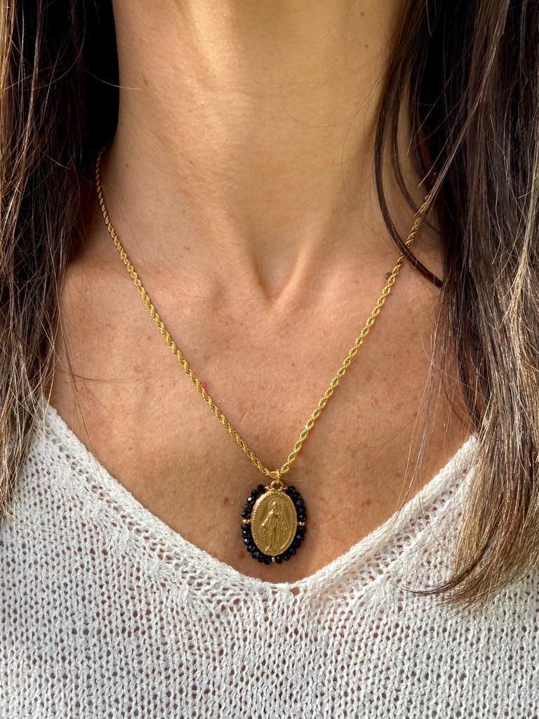 Colar dourado com pendente oval e pedras pretas em pescoço de pessoa com camisola branca