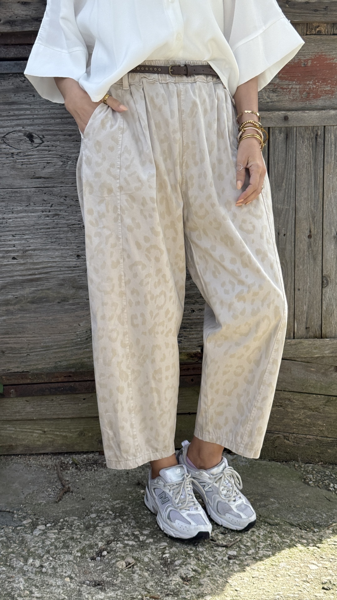 Calças bege com padrão de manchas, cinto castanho, sapatilhas brancas New Balance, camisa branca, pulseiras douradas, em cenário exterior com chão de pedra.