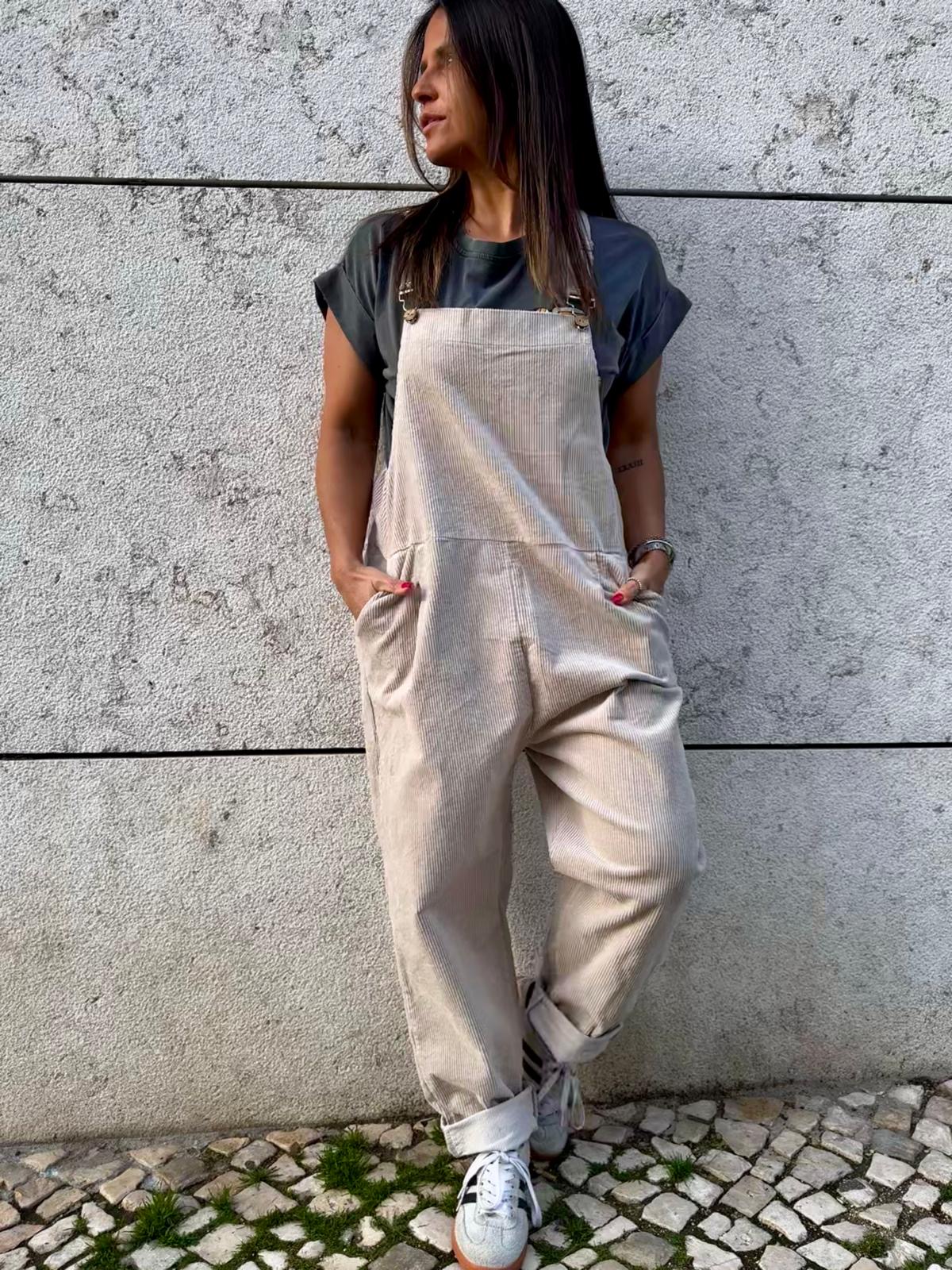 Mulher veste macacão bege com t-shirt cinza e sapatilhas brancas encostada a parede de cimento.