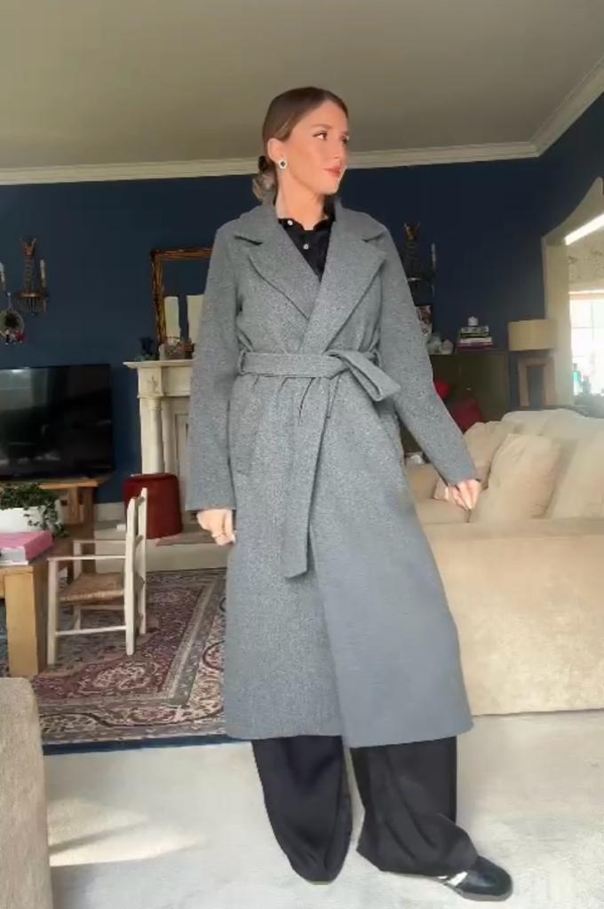 Mulher com casaco cinzento comprido com cinto numa sala de estar