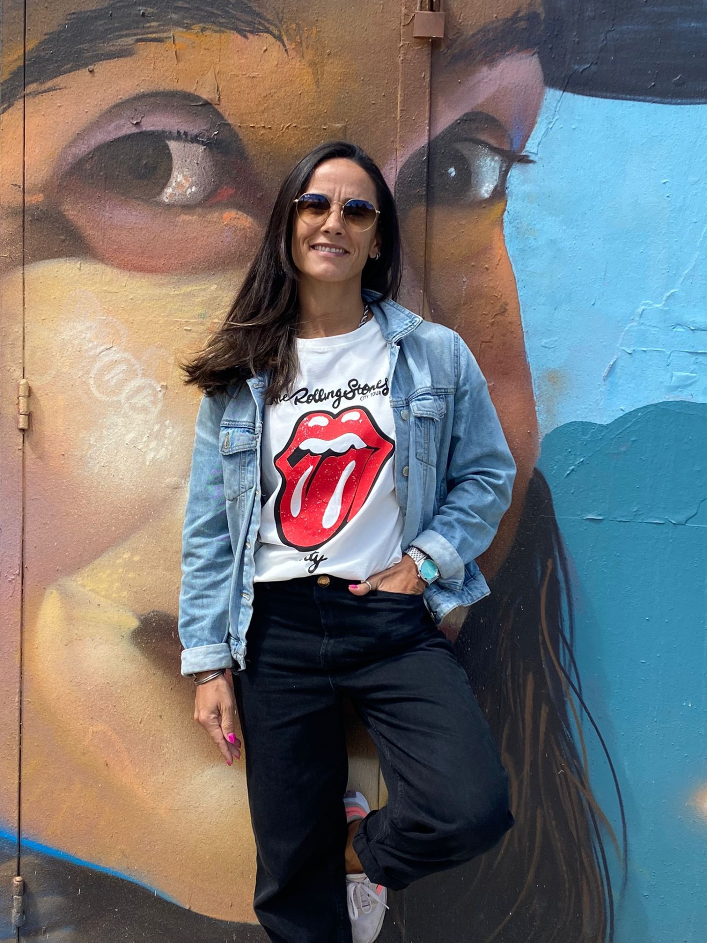 Mulher com t-shirt Rolling Stones e casaco azul encostada a parede com graffiti de rosto colorido.