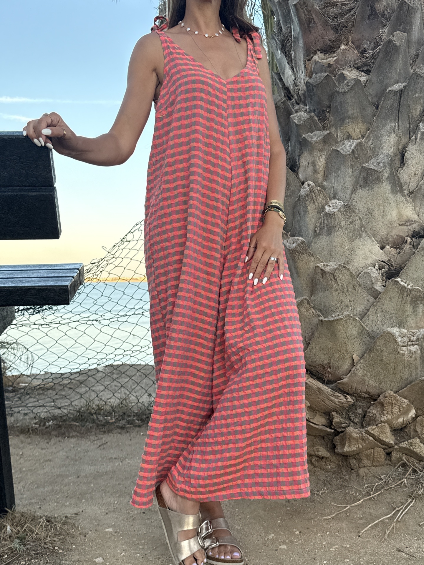 Vestido comprido vermelho e preto com padrão de quadrados e sandálias prateadas ao ar livre junto a uma árvore