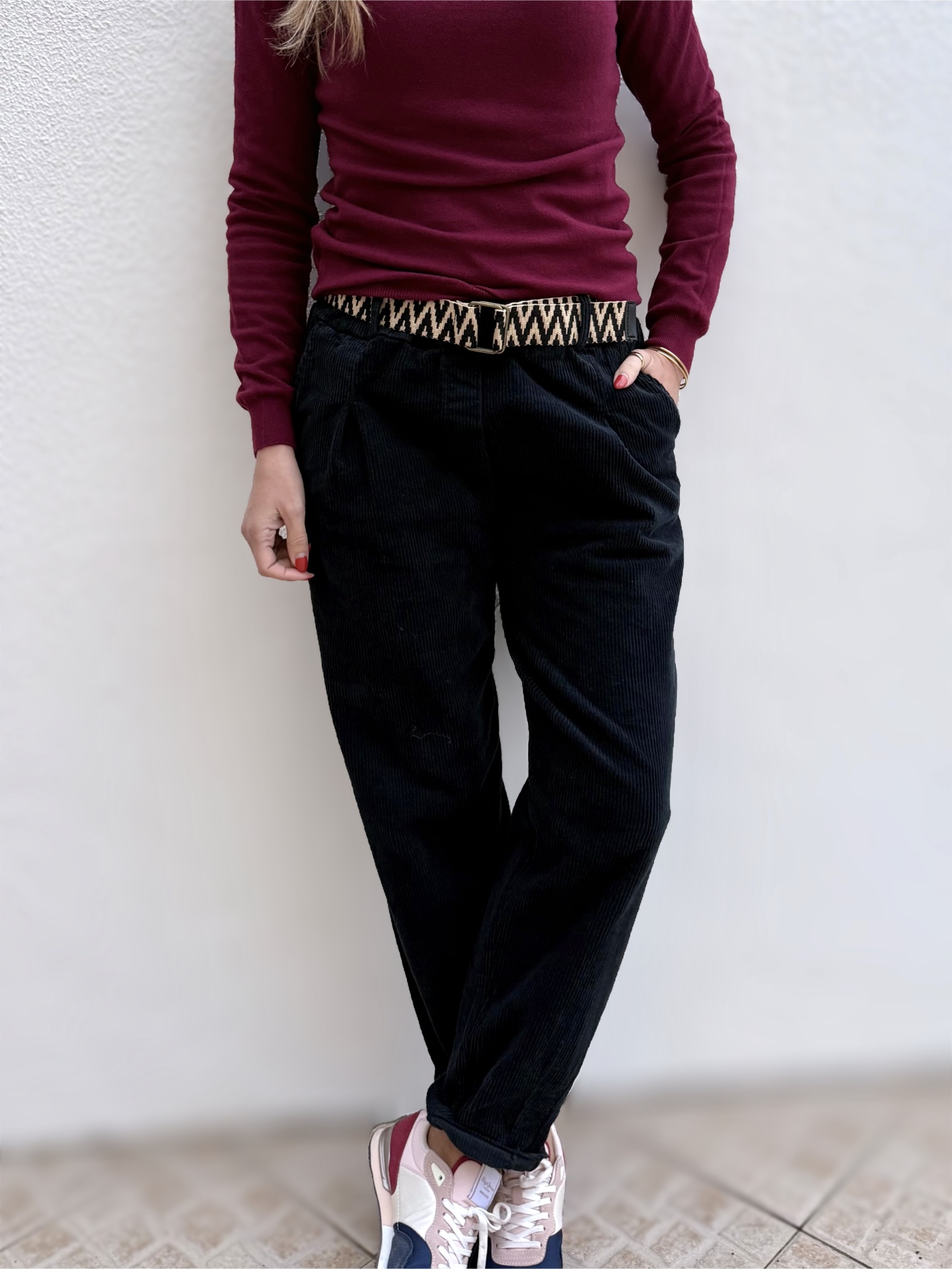 Pessoa com camisola vinho, calças pretas de veludo cotelê, cinto estampado e sapatilhas desportivas coloridas.