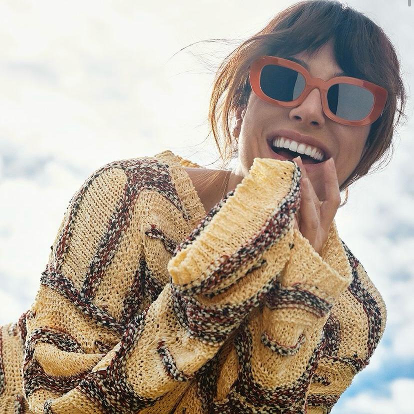 Pessoa com suéter de malha amarelo e castanho e óculos de sol castanhos sorrindo ao ar livre