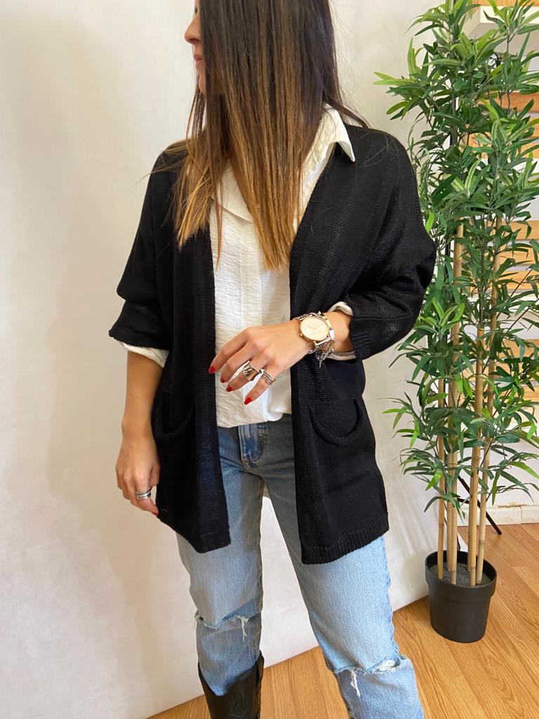 Mulher com cardigan preto, camisa branca, jeans rasgados e botas pretas ao lado de planta em vaso