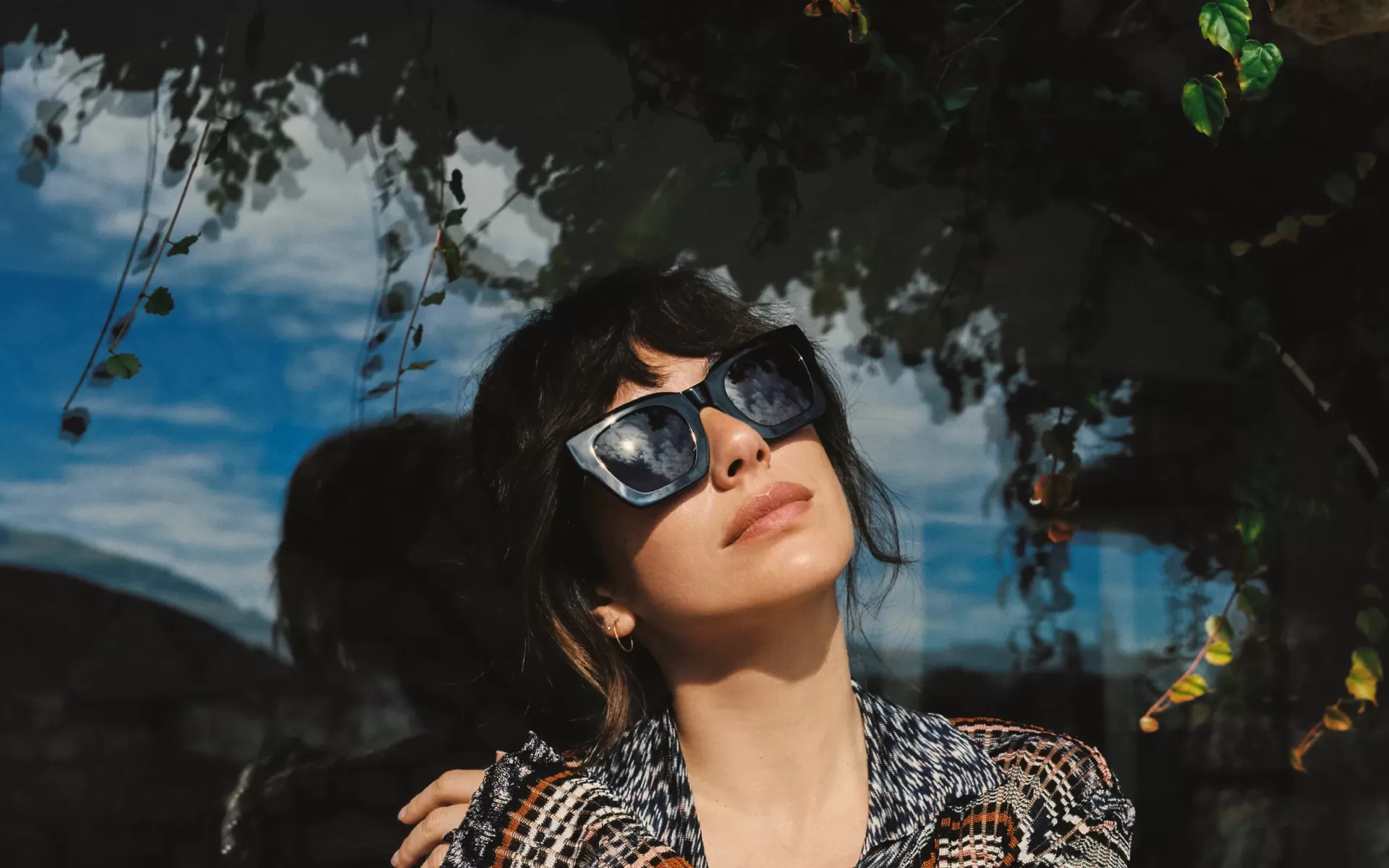 Mulher com óculos de sol pretos e casaco de padrão às riscas em fundo refletivo escuro