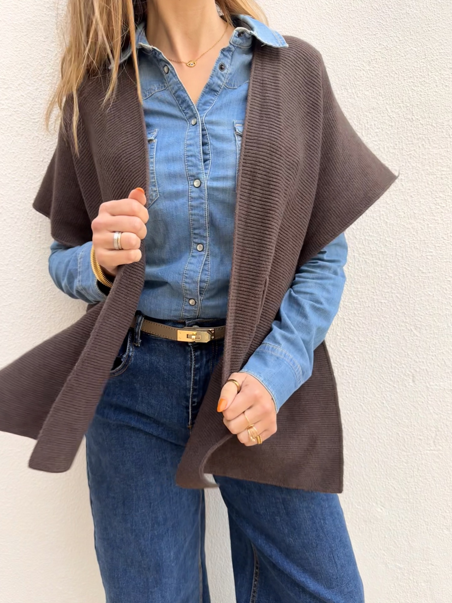 Modelo com camisa de ganga azul, colete castanho, jeans azul escuro e acessórios dourados em fundo branco.