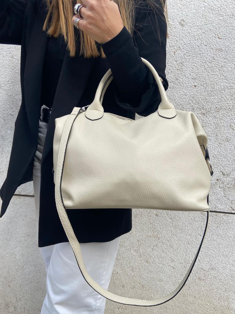 Mala de mão bege com alça longa, texto mostrando textura em couro, contra fundo de parede cinzenta