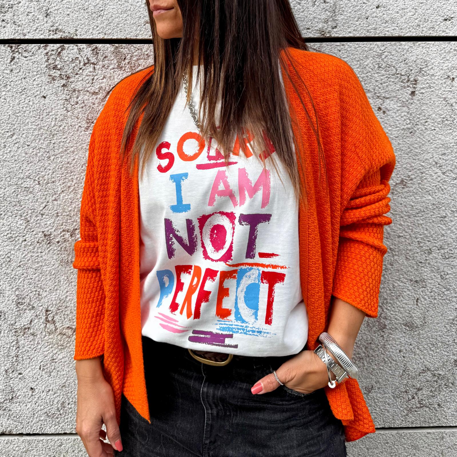 Mulher com camisola branca estampada e cardigan laranja junto a uma parede cinzenta