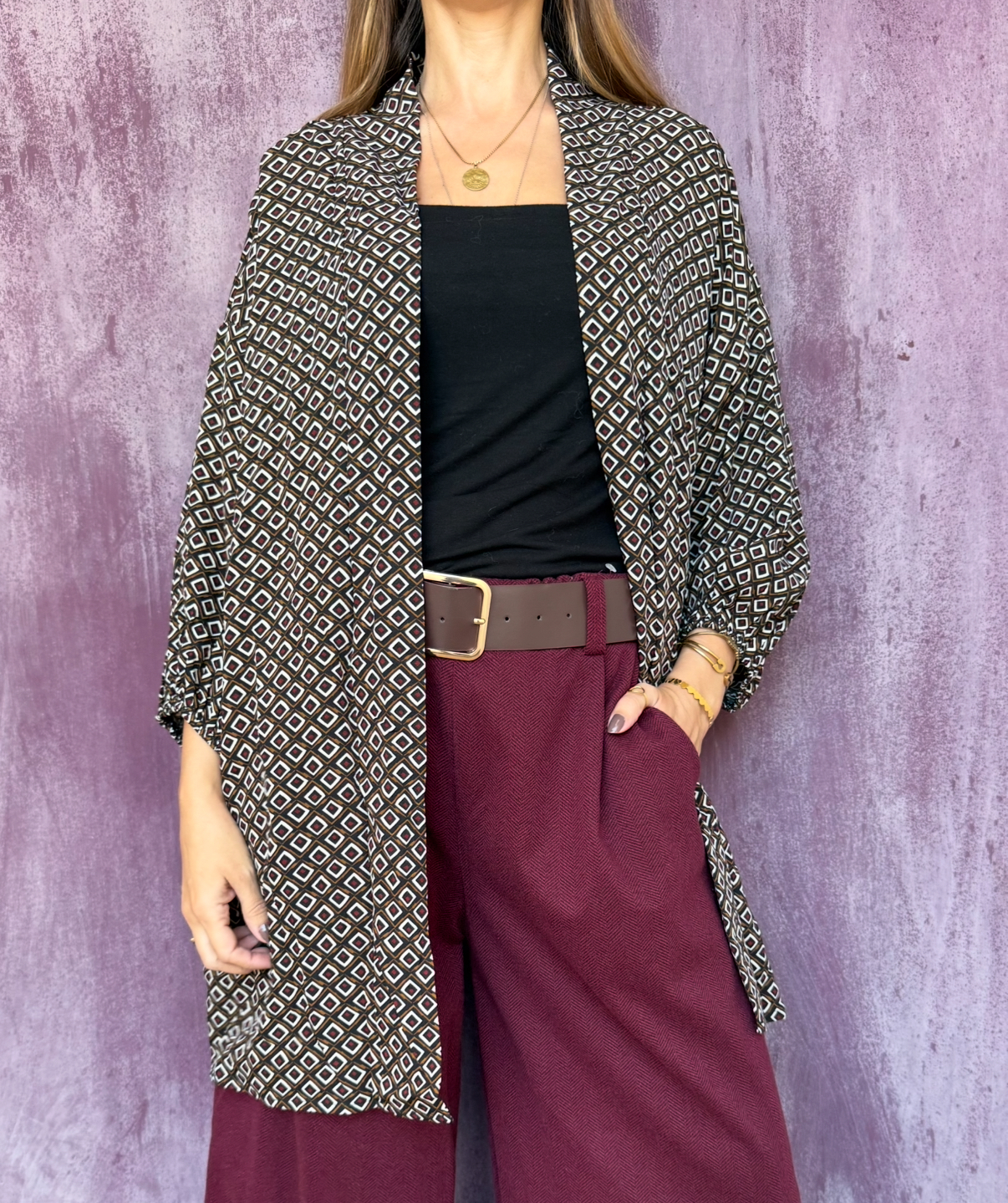 Pessoa veste kimono padrão geométrico castanho e branco, top preto e calças vinho com cinto castanho, fundo roxo.