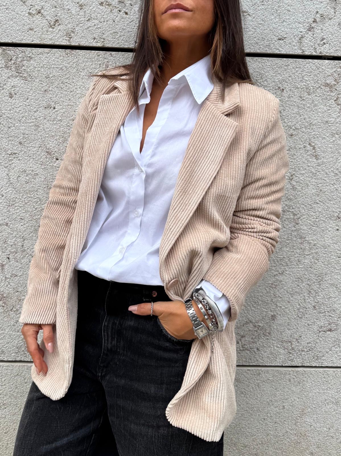 Mulher com casaco bege de veludo cotelê, camisa branca e calças pretas junto a parede cinza