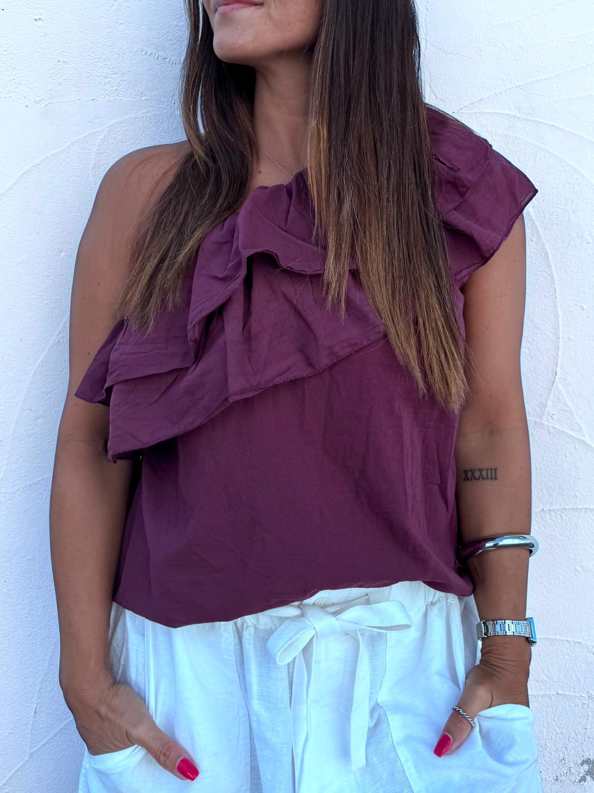 Blusa feminina roxa com folhos num ombro e calças brancas com cordão
