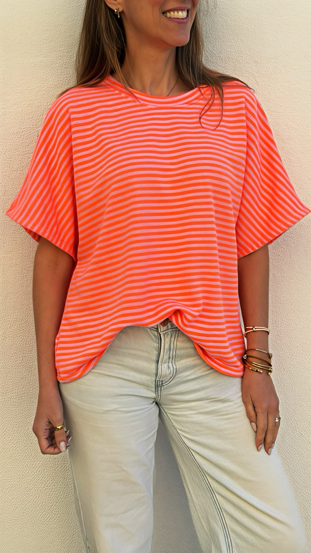Mulher com t-shirt laranja e rosa às riscas e calças claras junto a uma parede creme.