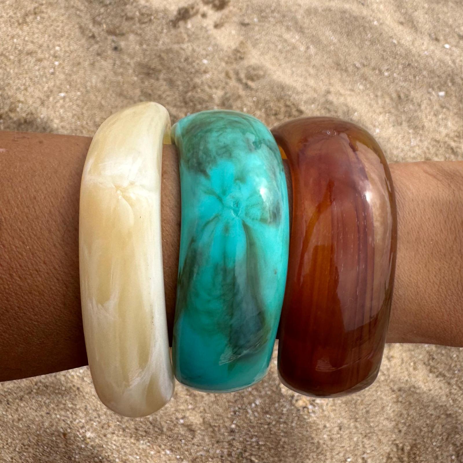 Três pulseiras largas de resina bege, turquesa e castanha num braço com areia ao fundo.