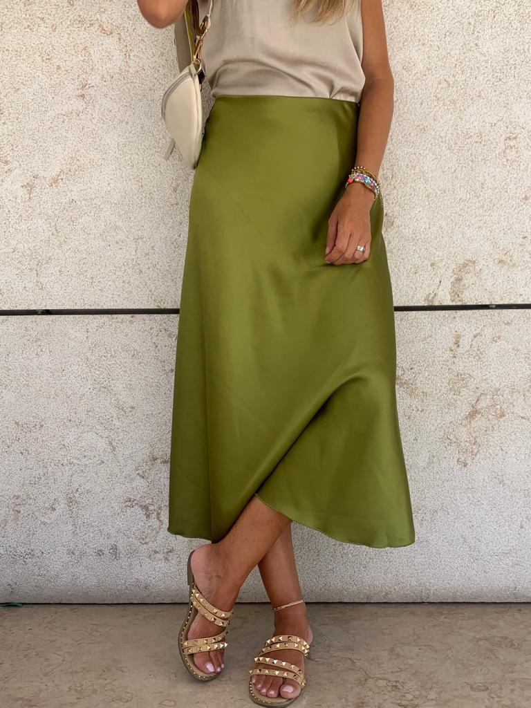 Saia verde oliva acetinada com corte assimétrico, sandálias baixinhas, top bege e mala pequena, em fundo de parede clara