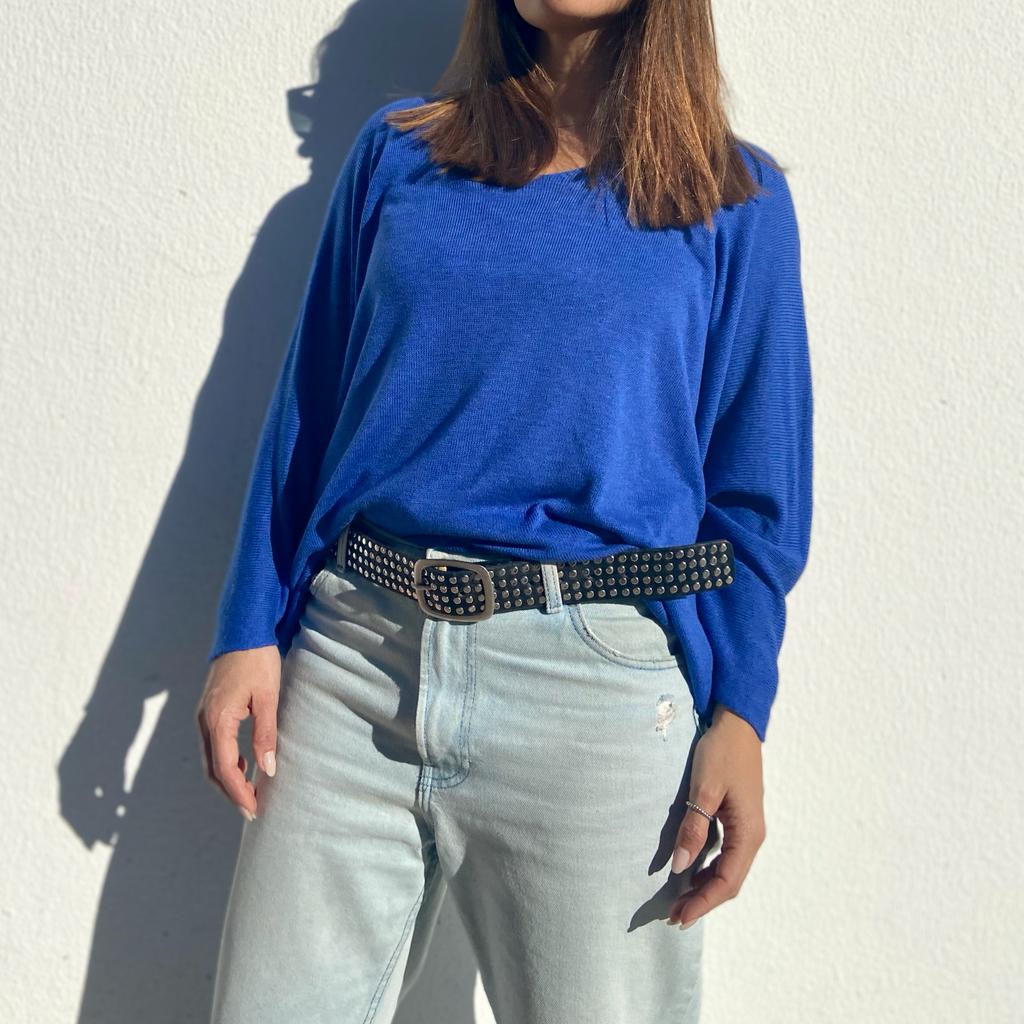 Pessoa com camisa azul de manga comprida, jeans claros e cinto preto com tachas contra parede branca.