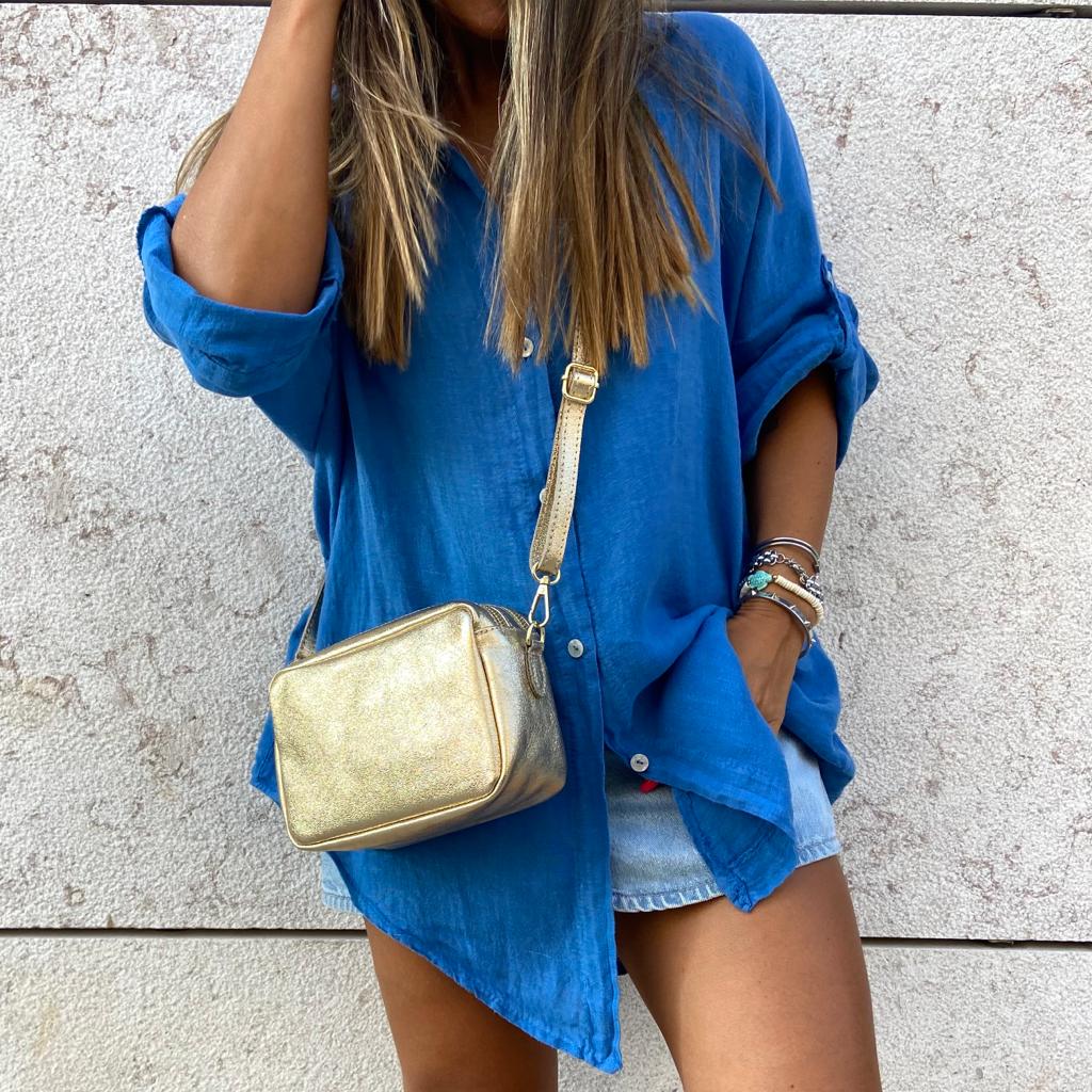 Pessoa com camisa azul, bolsa dourada, saia jeans curta junto a parede bege