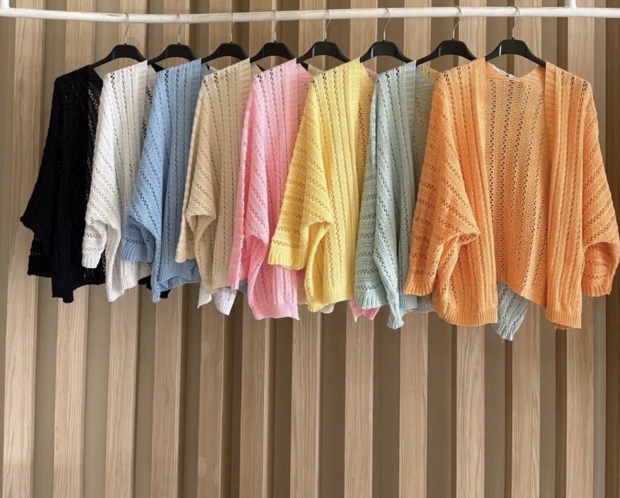 Oito casacos de malha coloridos pendurados em cabides contra painel de madeira clara.