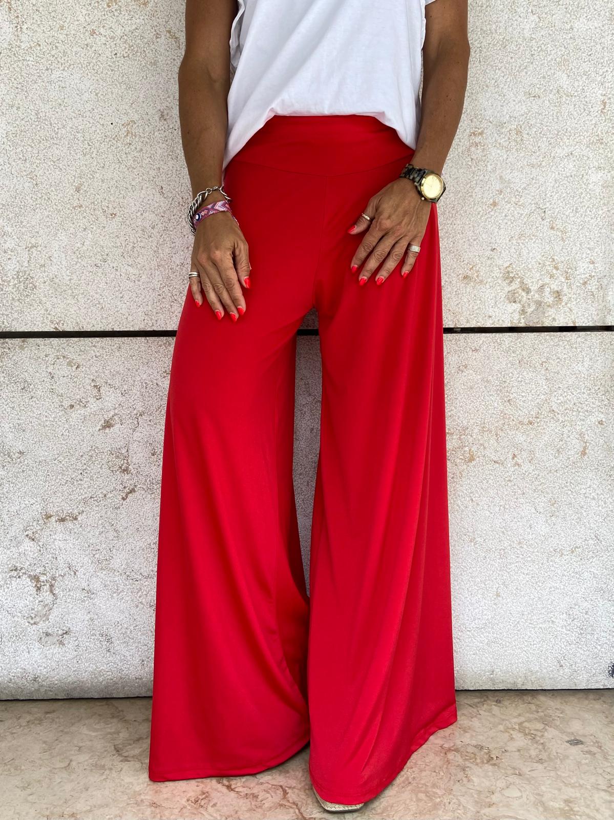 Pessoa usando calças largas vermelhas e t-shirt branca com parede de pedra ao fundo