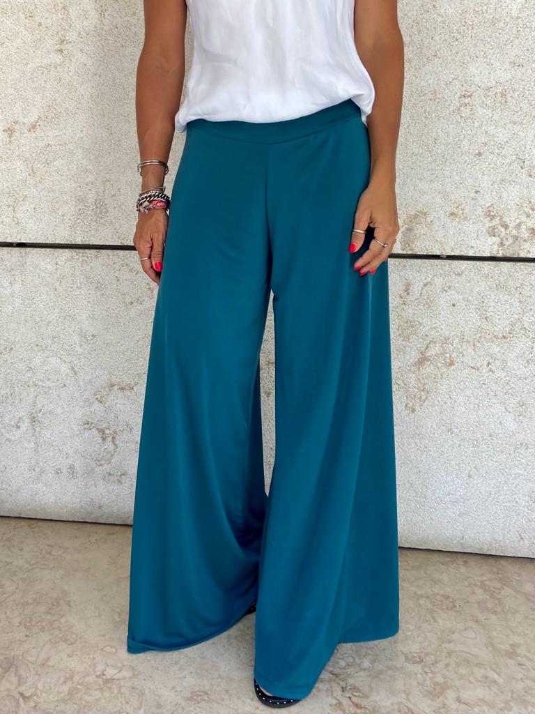 Calças largas femininas azul-petróleo com blusa branca em fundo de parede clara