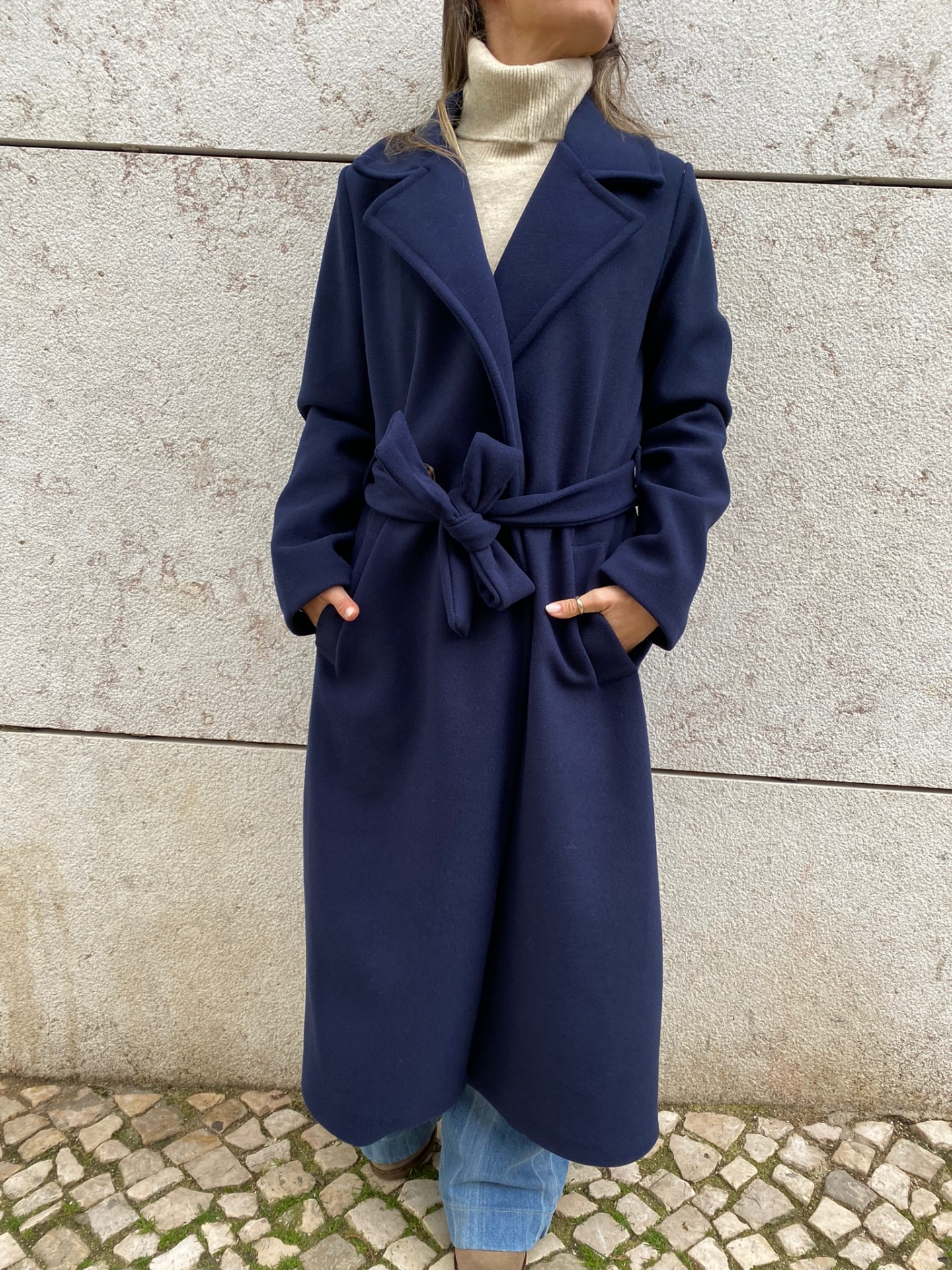 Casaco comprido azul escuro com cinto e bolsos, usado por pessoa com gola alta creme e calças azuis