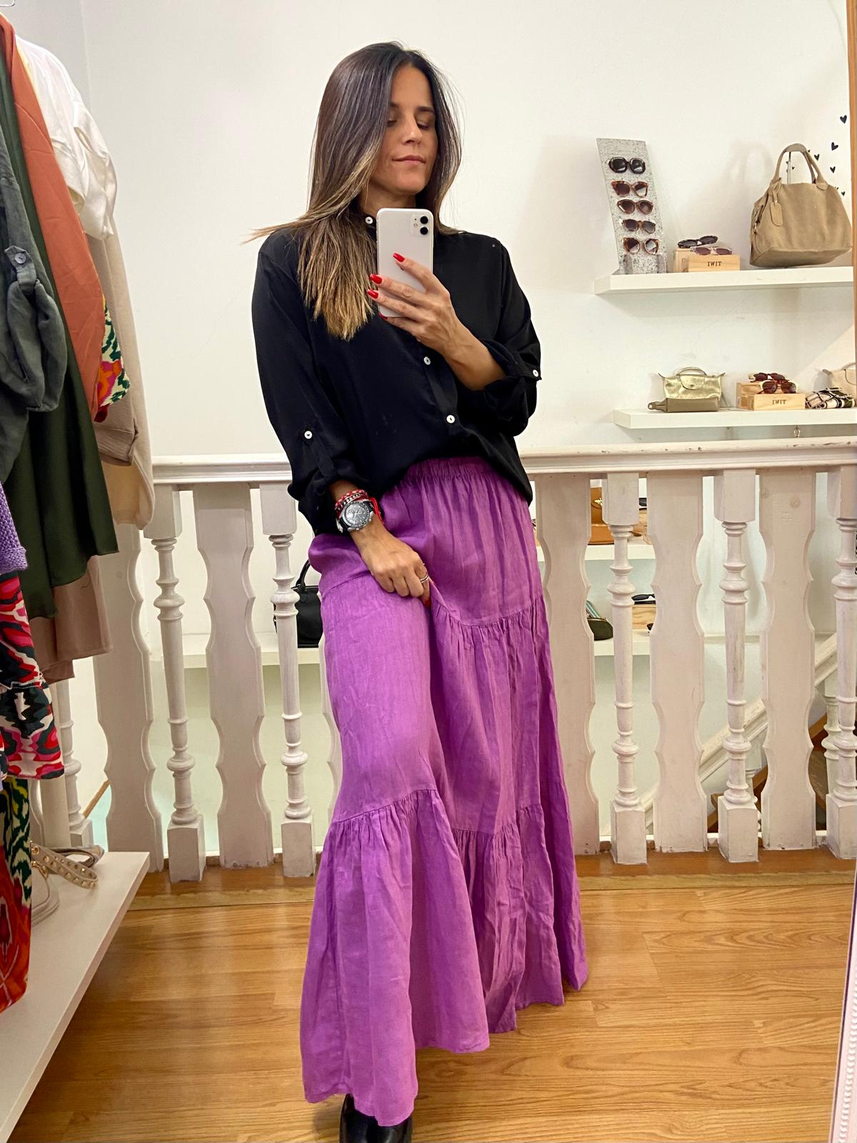 Mulher vestida com saia lilás comprida e blusa preta segura telemóvel branco em cenário interior com chão de madeira.