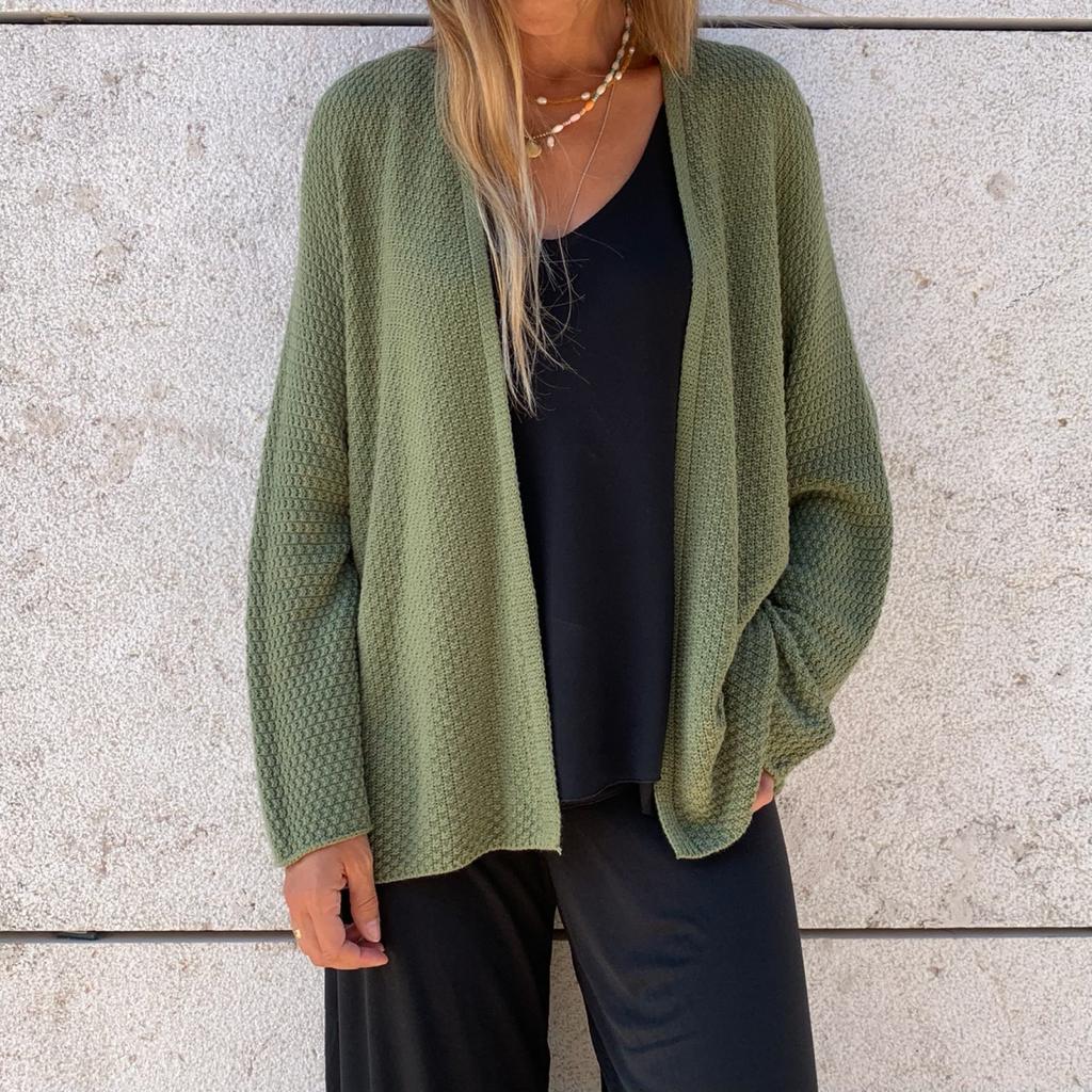 Casaco verde seco de malha, padrão texturizado, usado com roupa preta e fundo de parede clara